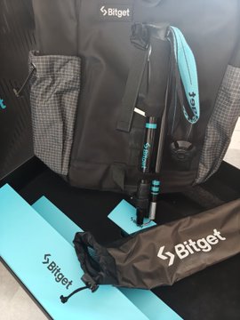 A black backpack and folded umbrella branded with "Bitget" are displayed alongside a teal blue mat, matching the post's description of stylish outdoor merchandise in black and Bitget's signature teal color scheme.