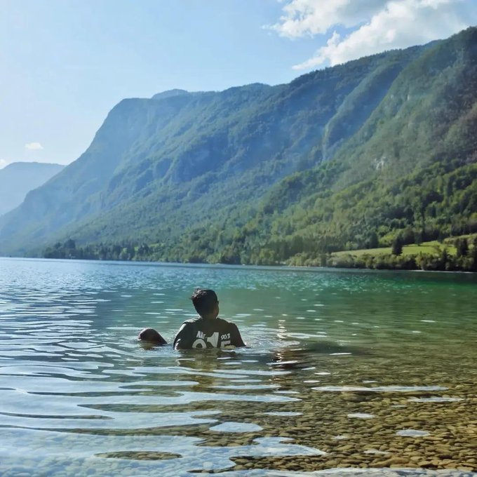 Two people swim in a clear lake surrounded by lush green mountains under a bright blue sky.