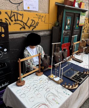 The image shows an outdoor market with a table displaying various items, including bracelets and eyeglasses. A person wearing a white shirt can be seen sitting at the table, possibly selling or showcasing these items. The surrounding area also features graffiti on the wall behind the table, adding to the urban atmosphere of the scene.