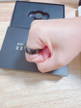 A person's hand wearing a silver smart ring on their finger, resting on a black box branded with the 'i' logo, likely the Ultrahuman Ring Air packaging.