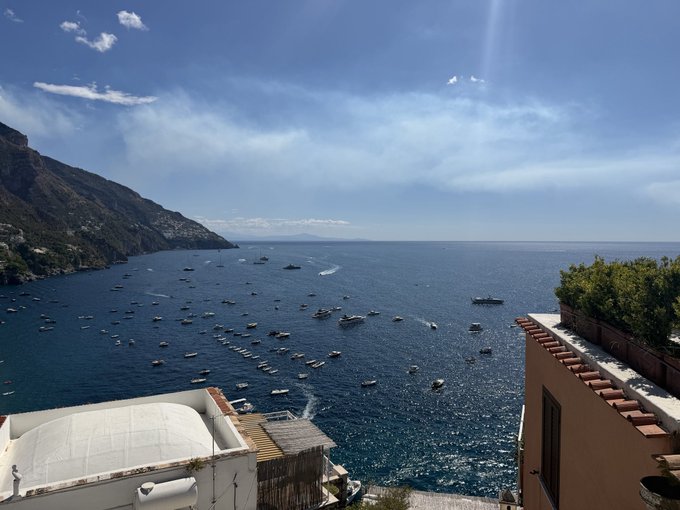 A scenic coastal view showing numerous boats anchored in blue waters along a mountainside, likely the Amalfi Coast or similar Mediterranean location.