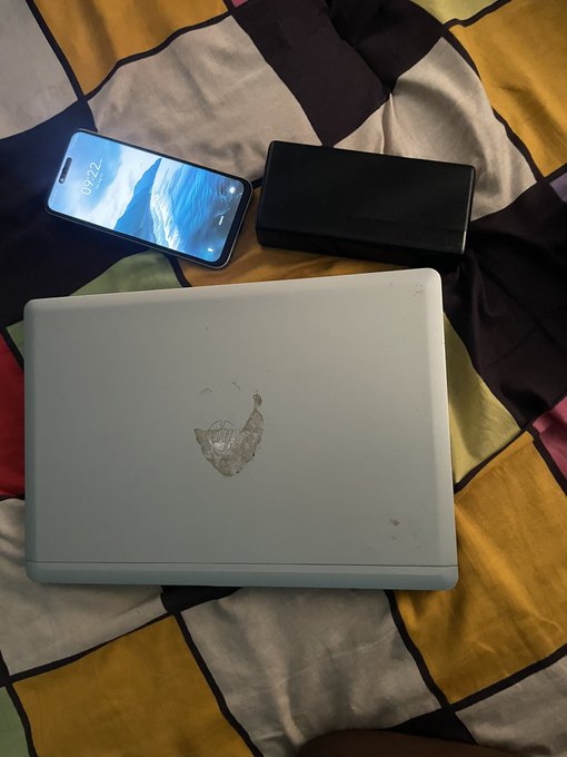 A silver laptop with a damaged/cracked logo area sits on a patterned bedsheet next to a smartphone displaying '09:22' and a black rectangular device.