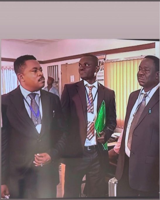 Photo of three men in business suits standing indoors, with the man in the middle looking sideways with a confused or skeptical expression while holding a green folder.