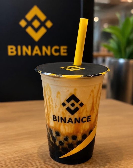A close-up photograph of a cup of bubble tea featuring the Binance cryptocurrency exchange logo, with a yellow straw and branded cup sleeve.