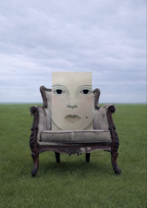 Surreal image of an ornate antique chair sitting on grass in a vast field, with a large pale mask-like face propped up where the chair's backrest would be.