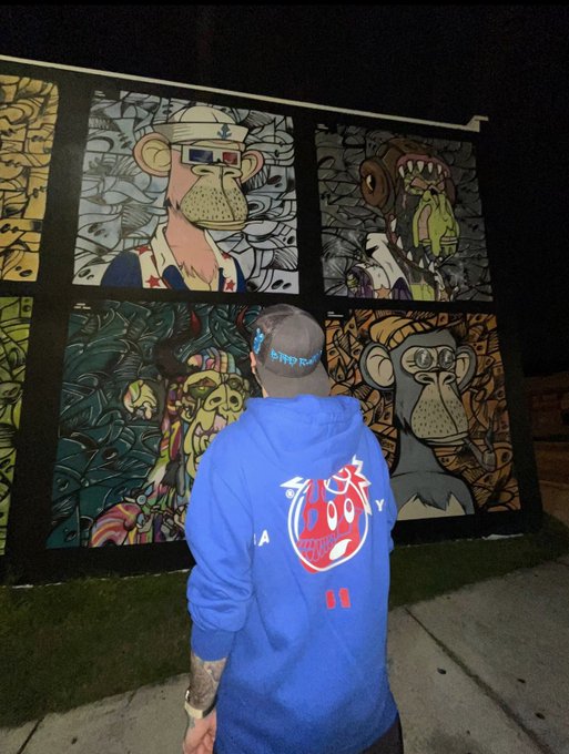 A man wearing a blue 'Bored Ape' hoodie stands with his back to the camera, looking at a wall display featuring Bored Ape Yacht Club NFT artwork including characters with 3D glasses and a zombie.