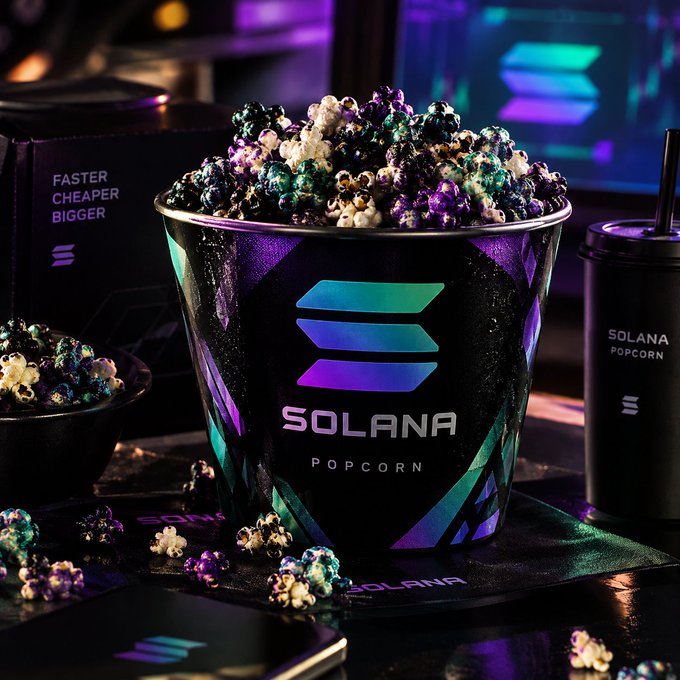 A large black bucket overflowing with iridescent purple, teal, and black popcorn is labeled "SOLANA POPCORN," accompanied by a matching cup and surrounding branded merchandise. The image playfully riffs on the "SOLGOGURU" sponsorship post by replacing the meme brand "GOGURU" with Solana's logo and offering crypto-themed popcorn instead.