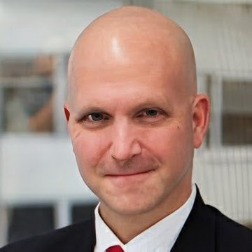 A professional headshot of a bald man in a black suit jacket, white shirt, and red tie, looking directly at the camera.