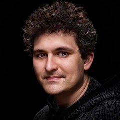 A portrait of a young man with curly dark hair smiling against a black background.