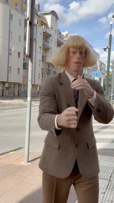 A person in a brown plaid jacket and blonde bob wig with a mustache stands on a city sidewalk, adjusting their tie.