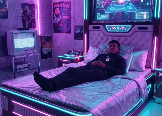 A person in dark clothing lies on a futuristic, neon-lit sci-fi bed illuminated with purple, pink, and green LED lights, with a retro TV showing static and posters on the concrete walls in the background.