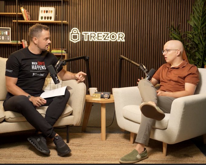 Two men sit in cream-colored armchairs having a discussion with microphones, in front of a wooden paneled wall featuring an illuminated "TREZOR" sign, while the post references the recording of a 1.5 hour conversation with Trezor's CTO discussing self-custody.