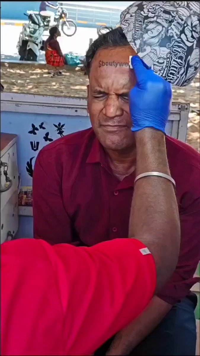 The image shows an older man with a bald head getting his hair shaved at a barber shop. A young boy is shaving another person's head while wearing blue gloves, possibly as part of a tattooing process or a medical procedure. The scene takes place in front of a store window, and there are people walking by outside the shop.