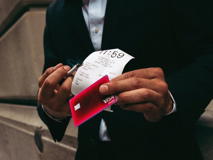 A person in a dark suit holds a Visa debit card and a receipt while writing with a pen, standing on stone steps.