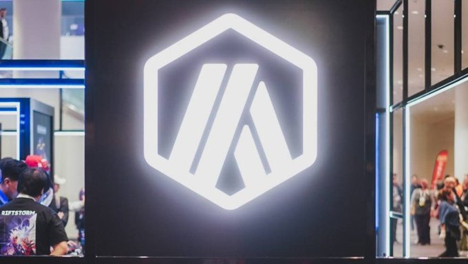An illuminated hexagonal logo is displayed on a wall at what appears to be a convention, with attendees visible in the background.
