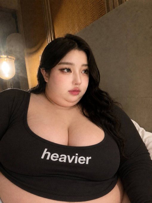A young woman poses in a dark gray long-sleeve shirt reading "heavier," referencing the post's playful quip about weight gain matching heavier pockets. The warm, moody lighting and intimate framing emphasize the shirt's message about her fuller figure.