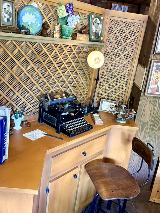 A vintage "ROYAL" typewriter sits on a wooden desk in a cozy home corner, surrounded by floral decor, framed family photos, and lattice paneling, with a wooden chair tucked neatly beneath the built-in cabinetry.