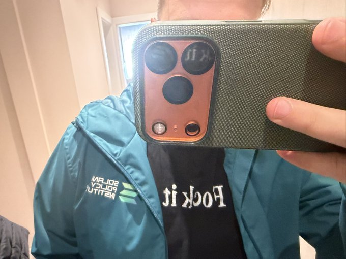 Mirror selfie of a person wearing a teal/green jacket with "AMITY SUBN SCREEN" text over a black "Fock it" t-shirt, holding a smartphone with a green textured cover and copper-colored camera module.