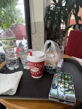 Casual photo on a black tray showing a bottle of water, a Holland Coffee cup with a red straw, a wrapped food item, a napkin, and an iPhone displaying a colorful grid of app icons.