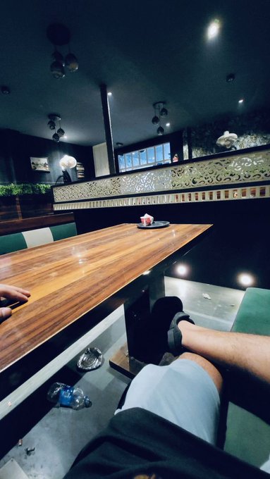 A dimly lit restaurant or lounge with a wooden communal table and decorative ornate divider, where a person in shorts is relaxing with their feet up while enjoying a quiet evening alone. The casual, contemplative atmosphere complements the post's tone of reflection amid the noisy Web3 space, as the author looks for projects like @quipnetwork and @TheARCTERMINAL that offer substance over hype.