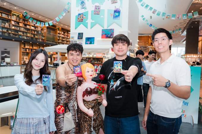 Four people pose together indoors holding colorful trading cards, standing beside a life-sized anime figurine in a bookstore or cafe setting decorated with banners.