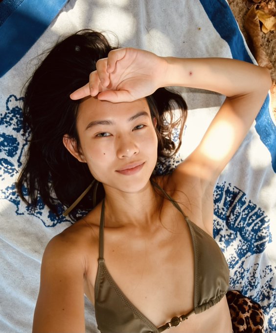A young woman with long dark hair wearing an olive green bikini top lies on a blue and white patterned towel, shielding her eyes from the sun with her hand.