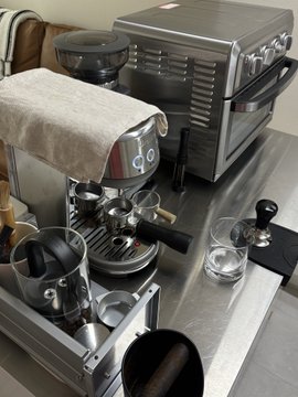 A stainless steel espresso machine sits on a kitchen counter alongside a portafilter, coffee grinder, and a small glass of water, all ready for use. The image perfectly captures the leisurely morning ritual of brewing coffee to shake off a hangover, with the home barista setup waiting patiently for its first pull of espresso.