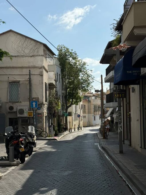 A sunlit Mediterranean street scene with parked scooters, narrow lanes, and warm-toned buildings under a blue sky.