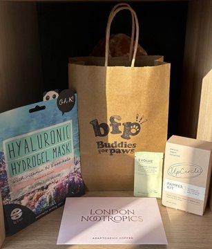 A curated gift bag for the Buddies for Paws Pilates event, featuring a bip Buddies for Paws tote alongside "OK!" Hyaluronic Hydrogel Mask, UpCircle coffee & paperback scrubs, and a London Kooptomics "Keep Going" card, with the sunset-lit London skyline peeking through the window behind the setup.