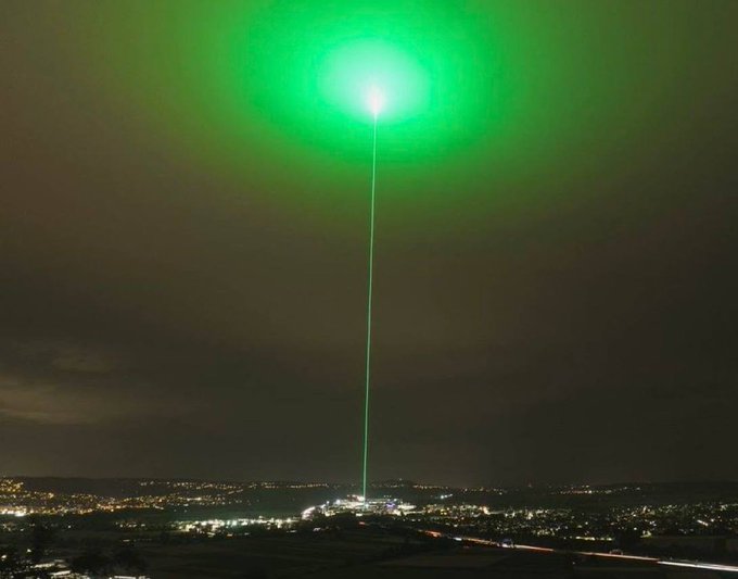 A vivid green laser beam shoots straight up into the night sky from an illuminated town, with a glowing green halo radiating overhead—visually mirroring the concept of a "god candle" shooting up rapidly like an unstoppable rally.

No visible text, numbers, tickers, or names appear in the image.