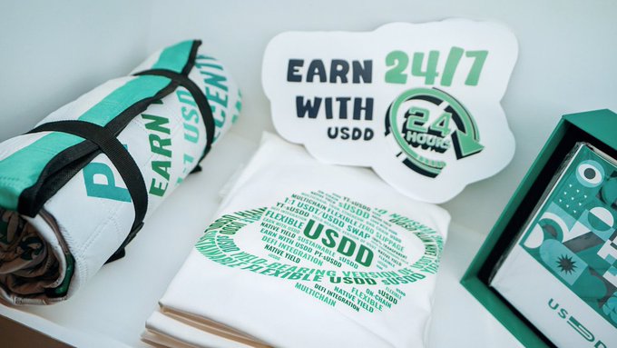 A promotional image for USDD stablecoin showing merchandise (t-shirt, sticker, and box) with text 'EARN 24/7 WITH USDD' and a 'FLEXIBLE' circular badge.