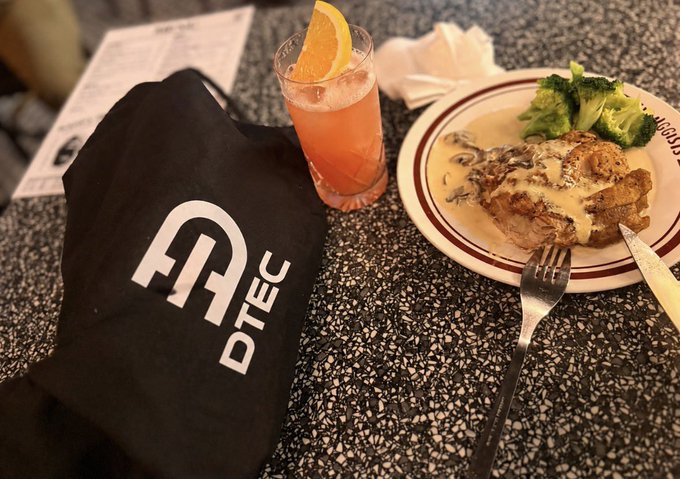 A meal on a speckled table consists of a plate with meat covered in mushroom sauce and broccoli, accompanied by a pink drink with an orange slice, a fork, and a black cloth bag bearing the logo 'DTEC'.