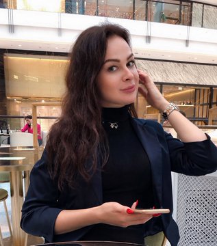 A portrait photo of a woman with long dark hair wearing a black turtleneck and blazer, seated in what appears to be a restaurant or lounge setting.