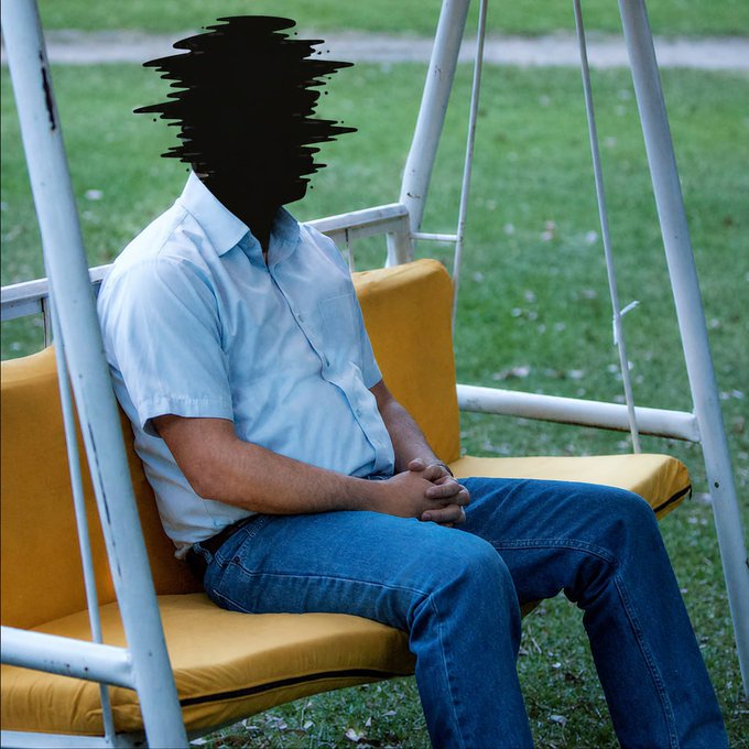 A man in a light blue short-sleeved shirt and blue jeans sitting on a yellow swing bench, with his face obscured by black scribbles, in a grassy outdoor setting.