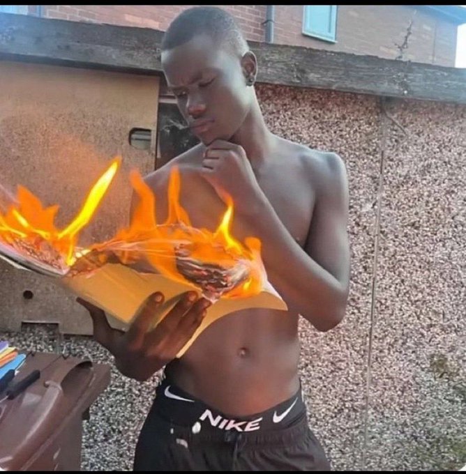 A shirtless young man wearing black Nike shorts stands outdoors looking thoughtfully at a book that has inexplicably caught fire in his hands.