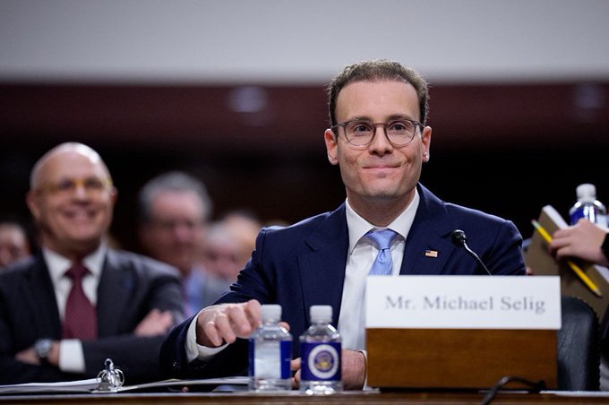 Michael Selig, identified by a nameplate reading 'Mr. Michael Selig,' sits at a formal hearing table in a dark blue suit and light blue tie, smiling slightly during what appears to be a U.S. congressional or regulatory proceeding.