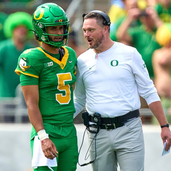 An Oregon Ducks college football scene showing quarterback #5 in a green and yellow uniform standing on the sideline talking with a coach wearing a white 'O' pullover and headset.