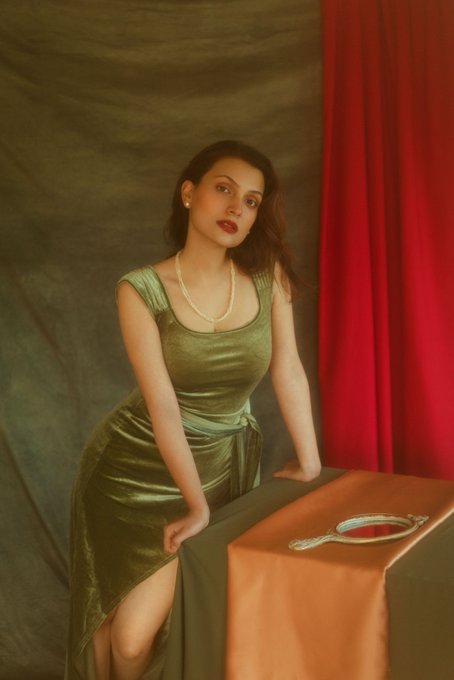 A woman in a green velvet dress and pearl necklace poses next to a table covered with orange and brown fabric, on which rests a hand mirror, with a red curtain backdrop.