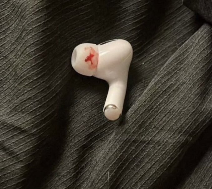 A single white wireless earbud lies on a dark textured leather surface, with a noticeable smear of fresh blood on the inner mesh where it contacts the ear.