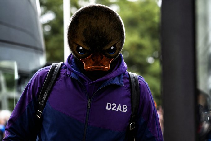 A person wearing a duck mask and a purple and blue jacket with 'D2AB' printed on it, carrying a backpack in an urban setting.