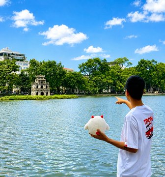 A person wearing a white t-shirt holds a stuffed animal while pointing across a lake toward Hoan Kiem Lake's Turtle Tower in Hanoi, Vietnam, under a bright blue sky.