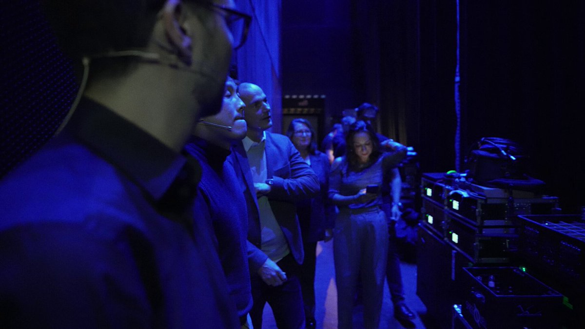 The image shows a group of people gathered together for an event at a theater or auditorium with blue lighting. The attendees are dressed formally, wearing suits and ties, indicating that they might be attending a special occasion or a formal event. One person is holding a microphone, suggesting that there may be some announcements or performances taking place during the gathering. The group appears to be engaged in conversation or observing something happening on stage, possibly related to the event's theme or purpose.