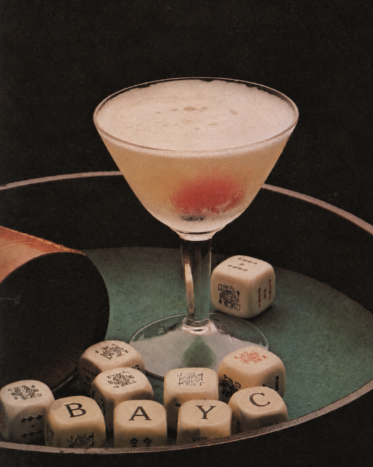 A cocktail glass with a cherry sits on a green surface surrounded by Scrabble tiles spelling "BAYC," with the accompanying post referencing the BAYC (Bored Ape Yacht Club) NFT collection.