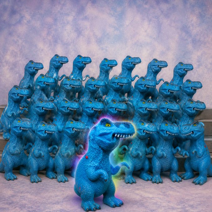 A group of identical blue T-Rex figurines arranged in rows, with one highlighted in the center surrounded by a glowing rainbow aura, symbolizing the original FEFER community standing together as "the old herd" that will not die.