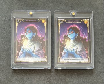 Two identical collectible cards in protective plastic sleeves featuring artwork of a figure with glowing energy, placed side by side on a gray surface.