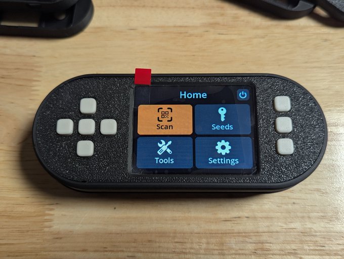 A SeedSigner Bitcoin hardware wallet device resting on a wooden surface, displaying its home screen menu with options for "Scan," "Seeds," "Tools," and "Settings," along with physical navigation buttons and a power button. The image showcases the portable, cable-free design of the device, complementing the post's appreciation for its battery-powered convenience and ease of use in collaborative Bitcoin security setups.