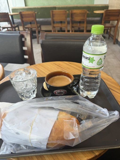 A tray on a wooden café table containing a wrapped bread item, a glass of ice water, a small ceramic cup of espresso with 'DEUTZ' branding, and a bottle of 'desani' water.