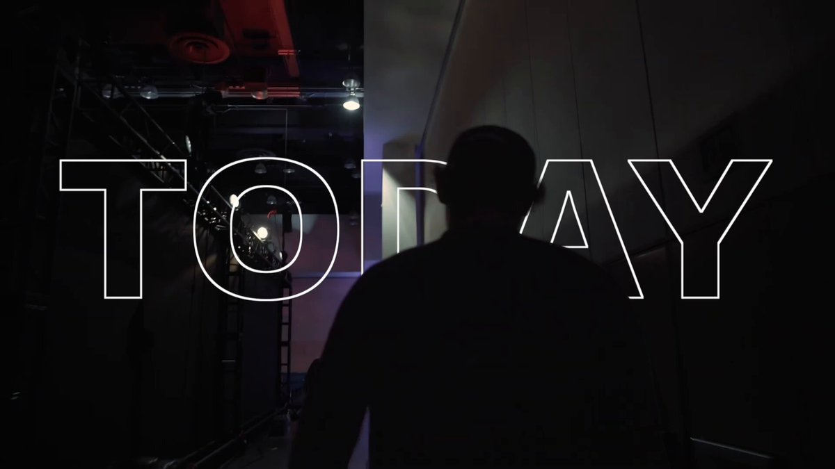The image features a man standing in front of a wall with the word "Today" written above him. The man appears to be looking at something off-camera, possibly an interesting object or event. The background seems dark, which contrasts with the white text on the wall and emphasizes the presence of the man.