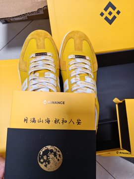 A pair of yellow sneakers branded with the "BINANCE" logo sits atop a yellow Binance box featuring Chinese characters that read "月满山海 敬和人堂" (Moon over mountains and seas, honoring harmony), tying into the user's Mid-Autumn Festival Binance gift unboxing.
