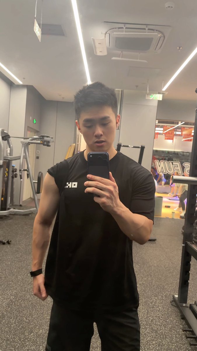A young man takes a mirror selfie in a gym while wearing a black "OKX" t-shirt, reflecting the OKX merch mentioned in his caption about his daily routine of working out, trading crypto, vibecoding, and playing soccer.